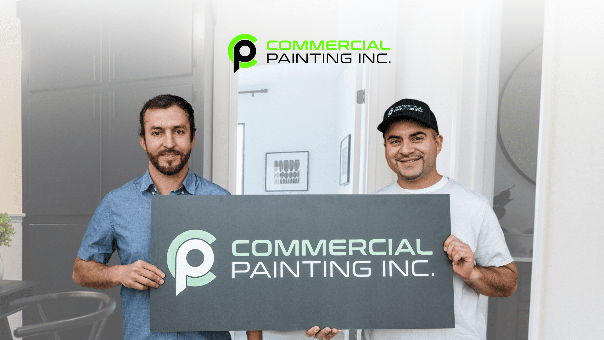 Two men holding a sign for Commercial Painting Inc. stand in front of a white refrigerator. One man wears a blue shirt and the other a white t-shirt and a baseball cap.