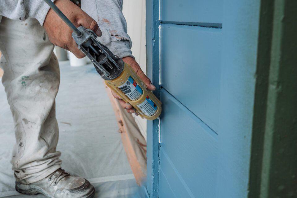 A man is applying silicone sealant to a blue wall.
