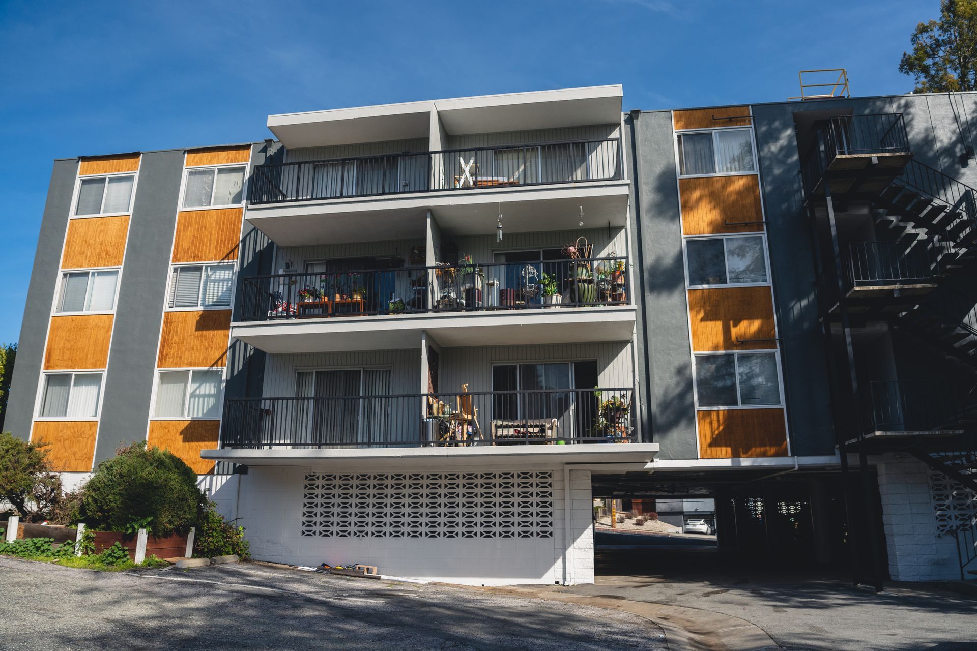 Multi-story apartment building with grey siding, orange accents, and balconies under a blue sky. Ground level has parking.