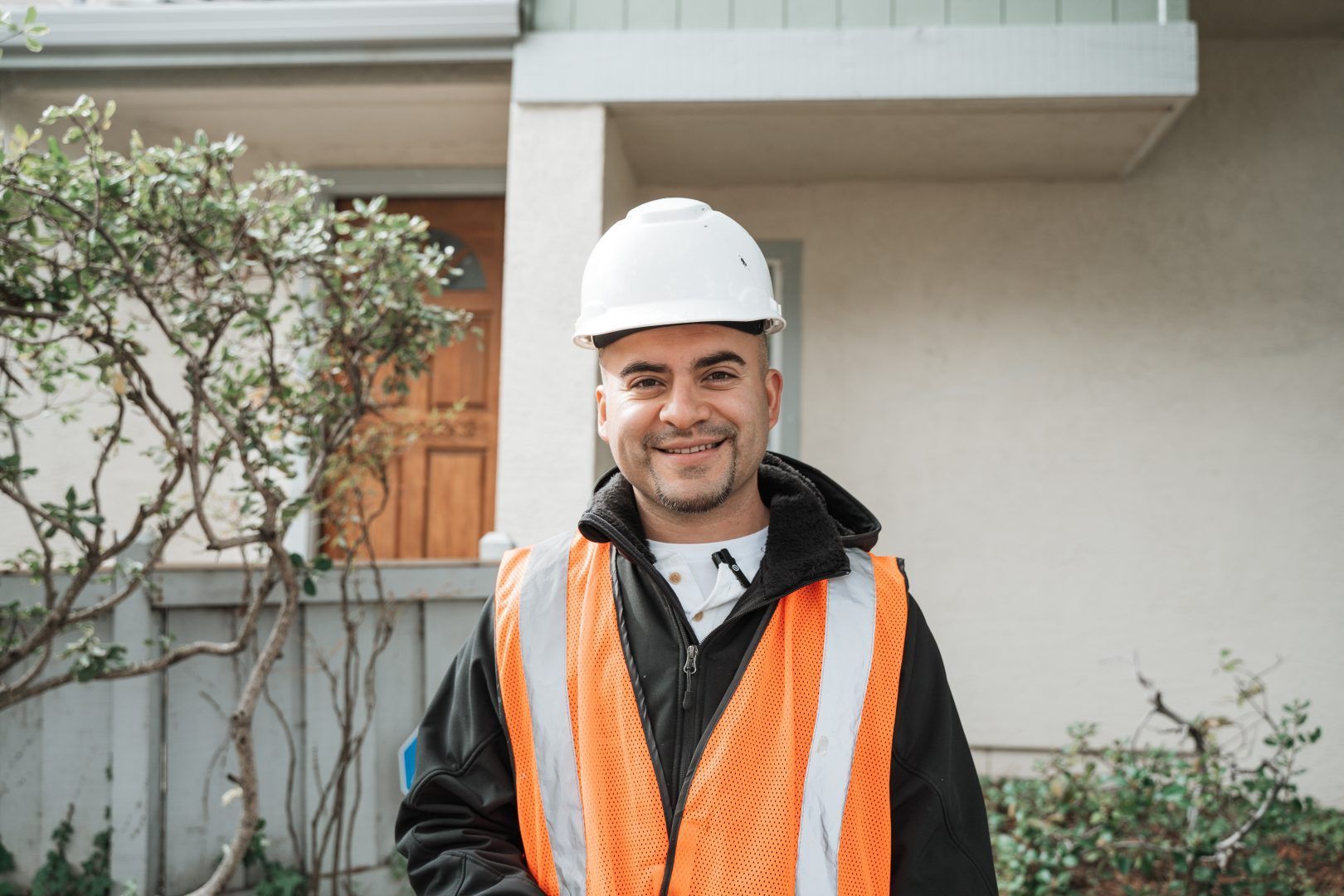 A man wearing a hard hat and an orange vest is standing in front of a house.