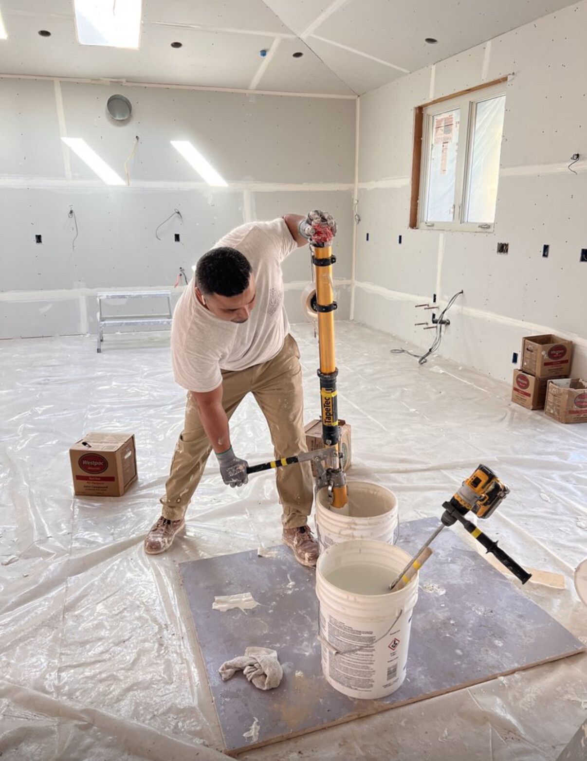 Man mixing joint compound in a construction setting. He's wearing a white shirt and khaki pants. White buckets and compound are visible.