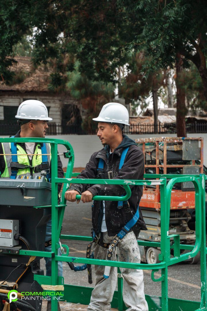 Two construction workers wearing hard hats are standing in a green cage.