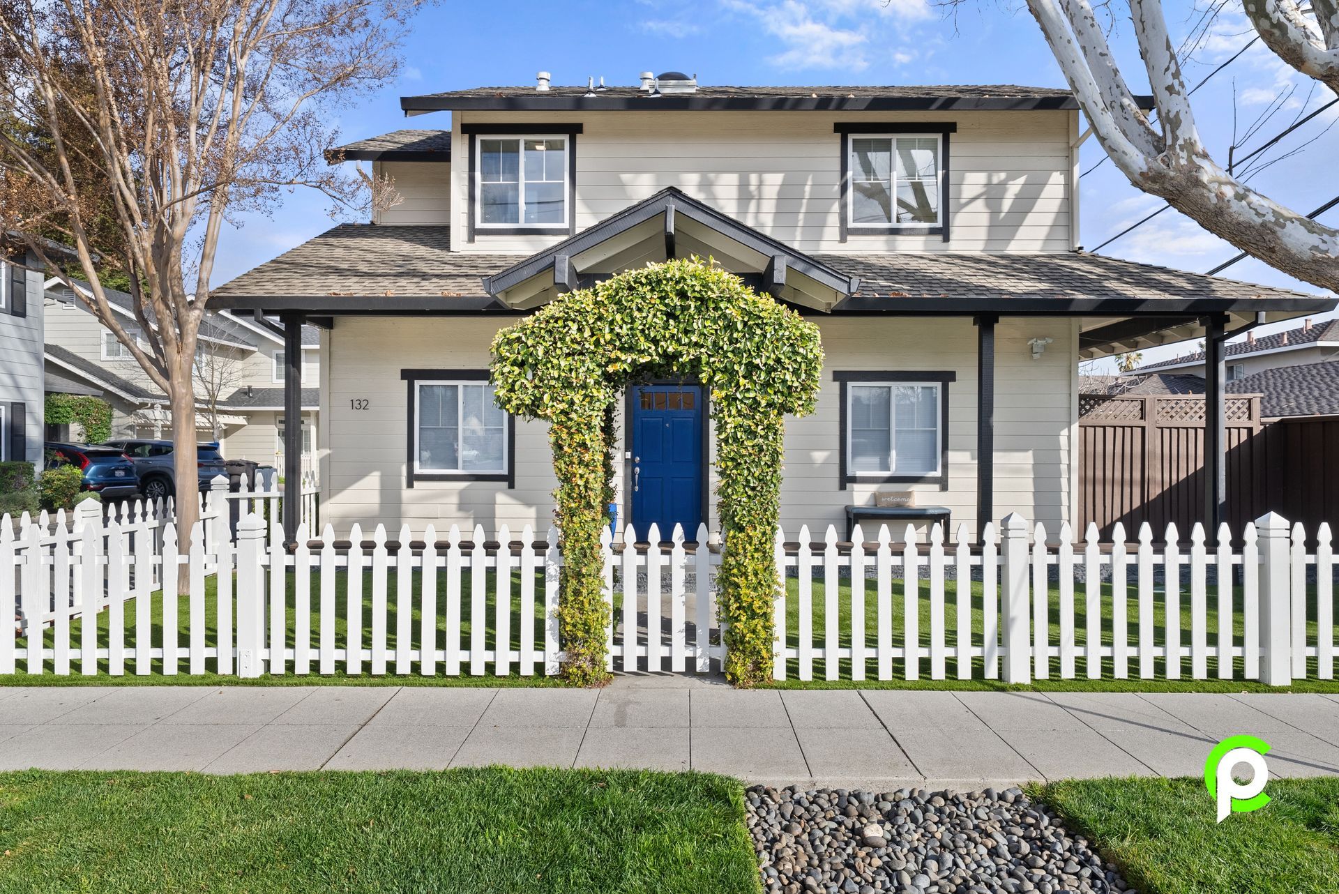 Two-story house with a white picket fence and a blue door, framed by greenery, under a blue sky.