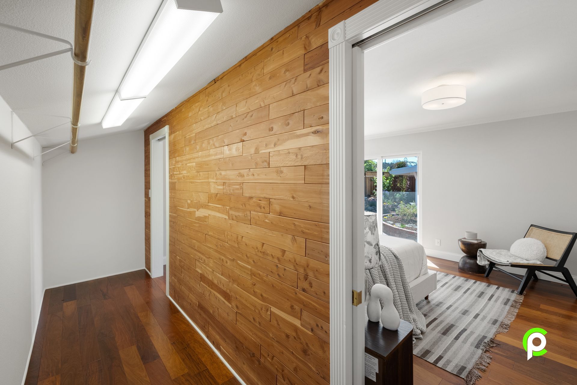 Hallway with wood-paneled wall leading to a bedroom with a bed, chair, and outdoor view.