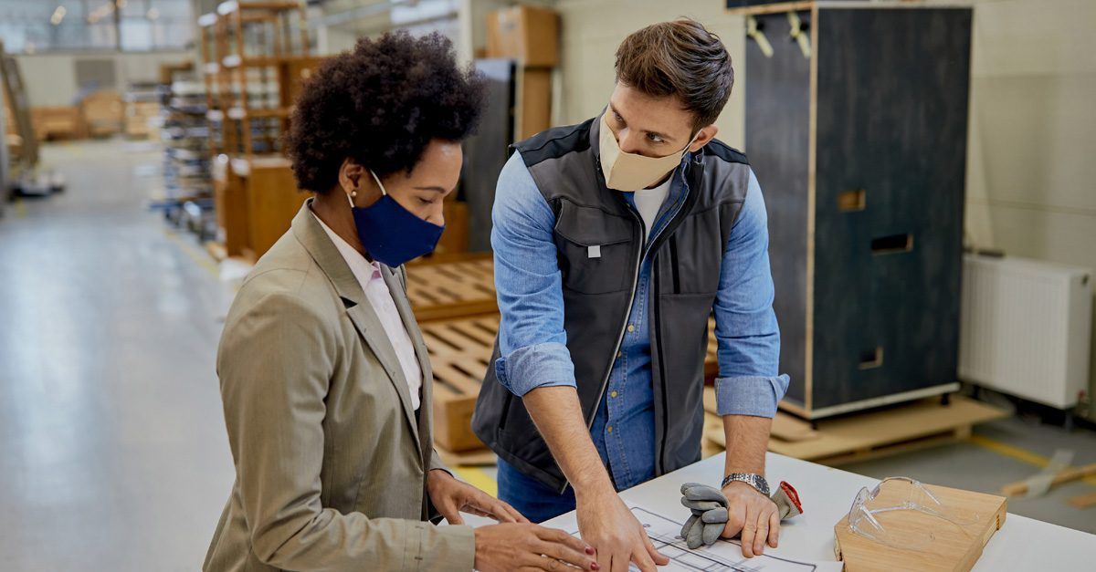 A man and a woman wearing face masks are looking at a blueprint in a factory.