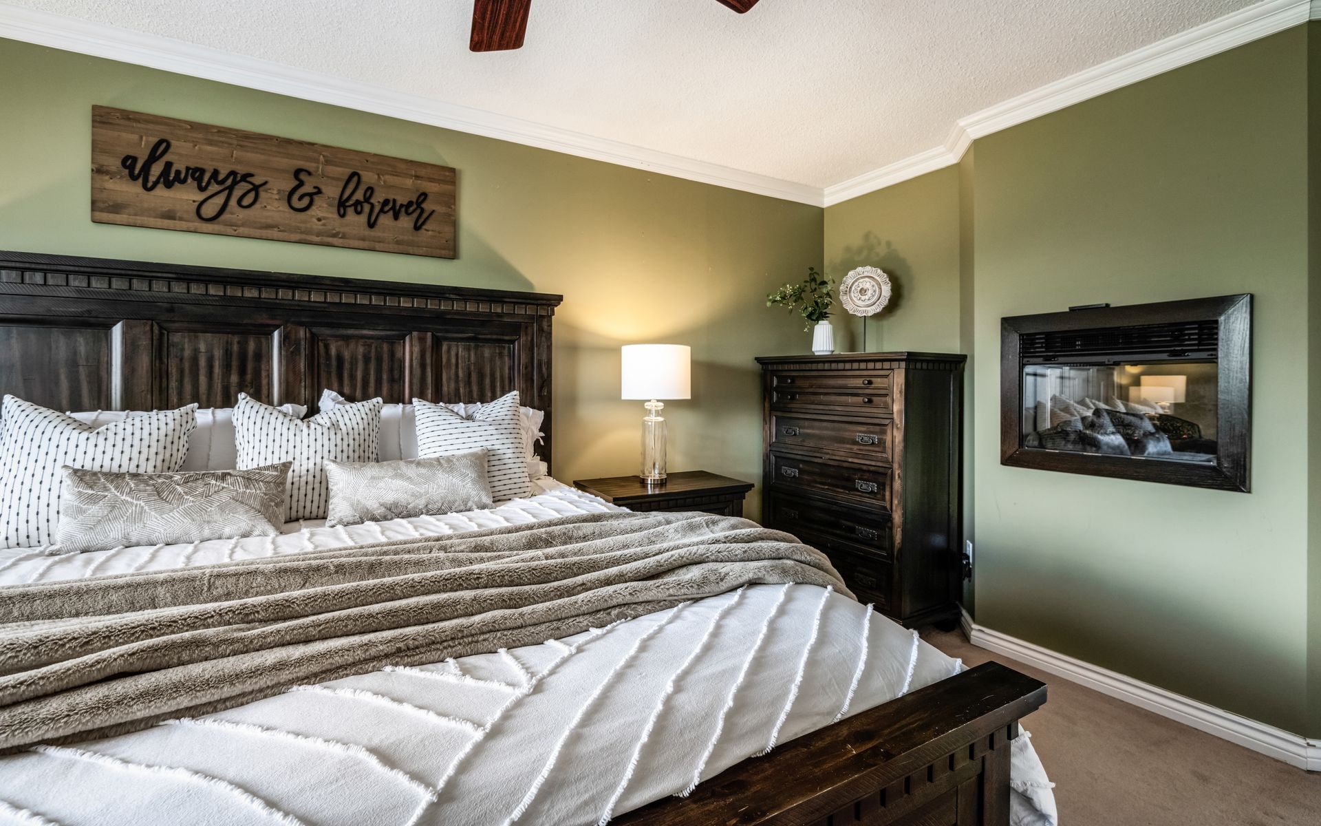 A bedroom with a king size bed , dresser , lamp and fireplace.