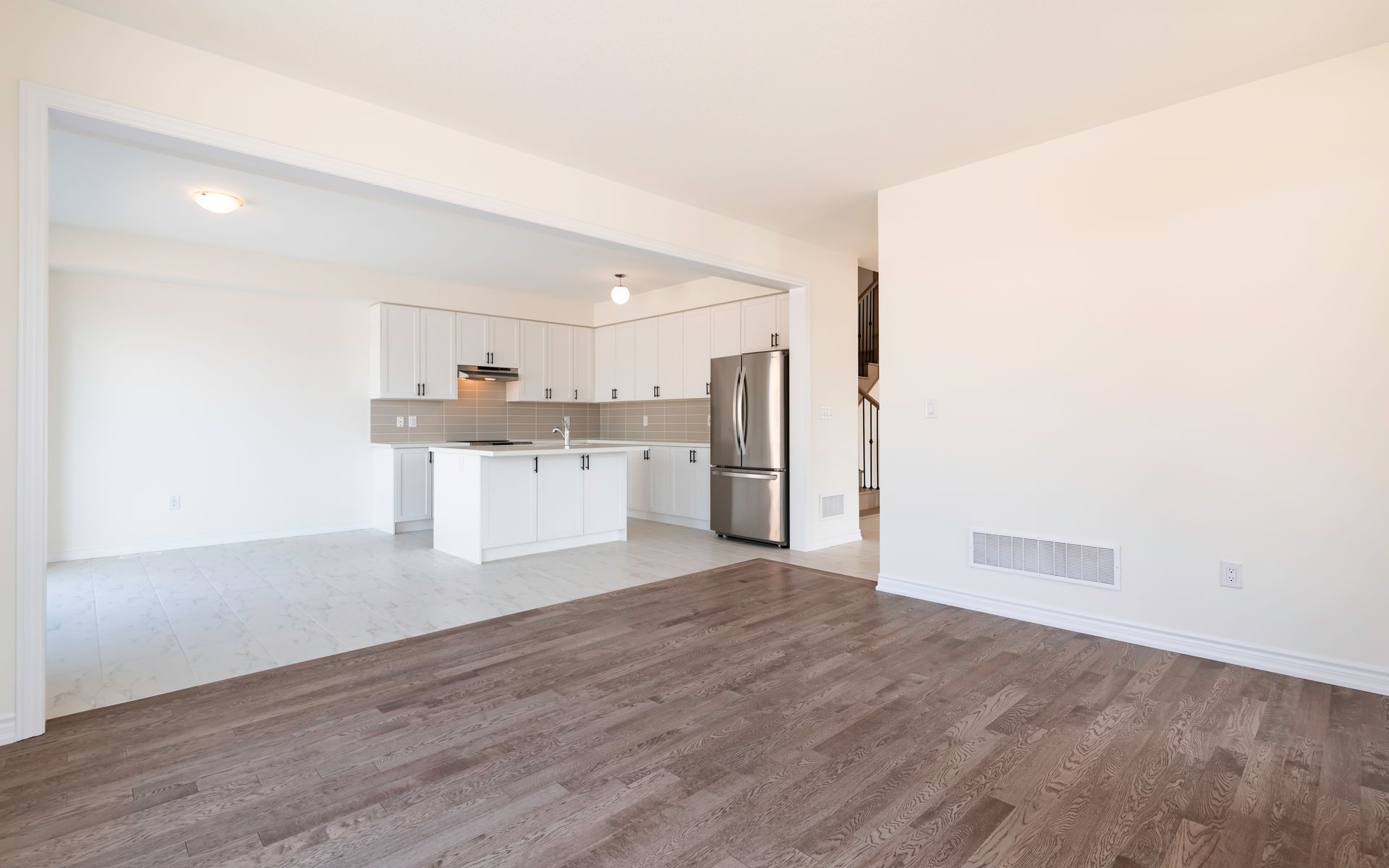 An empty living room with hardwood floors and a kitchen in the background.