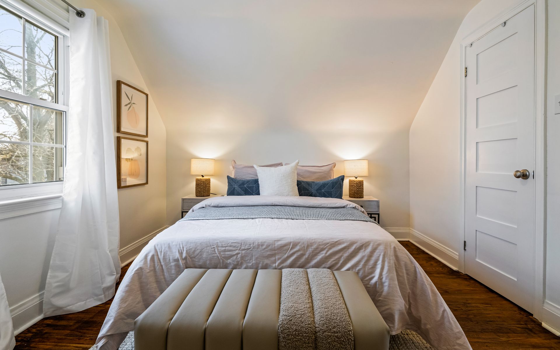 A bedroom with a bed , ottoman , lamps and a window.