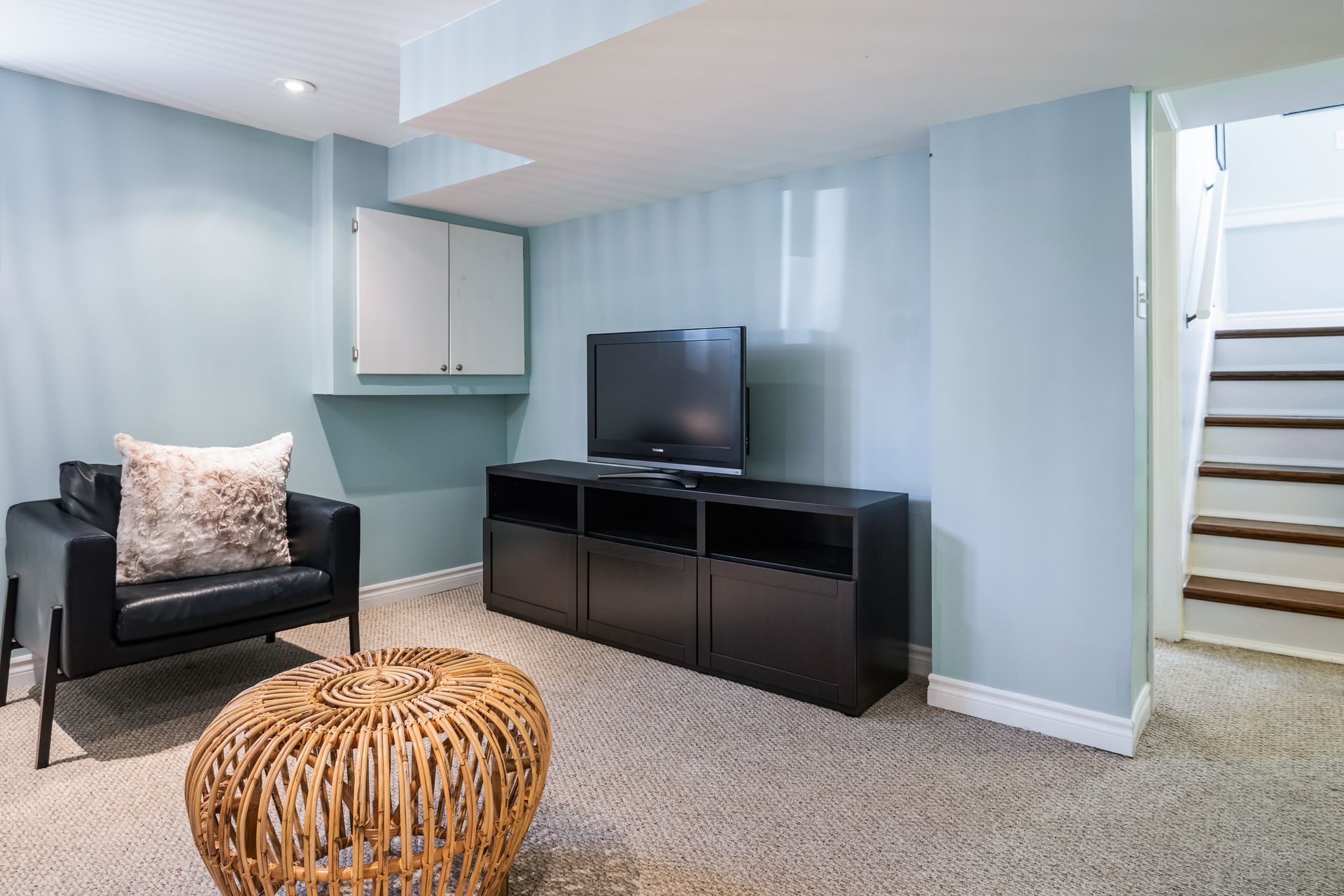 A living room with a chair , ottoman , television and stairs.