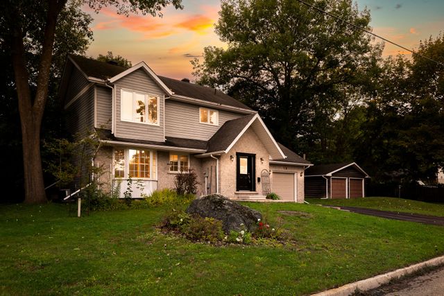 Two-story house with lit windows at sunset, surrounded by trees and a green lawn.