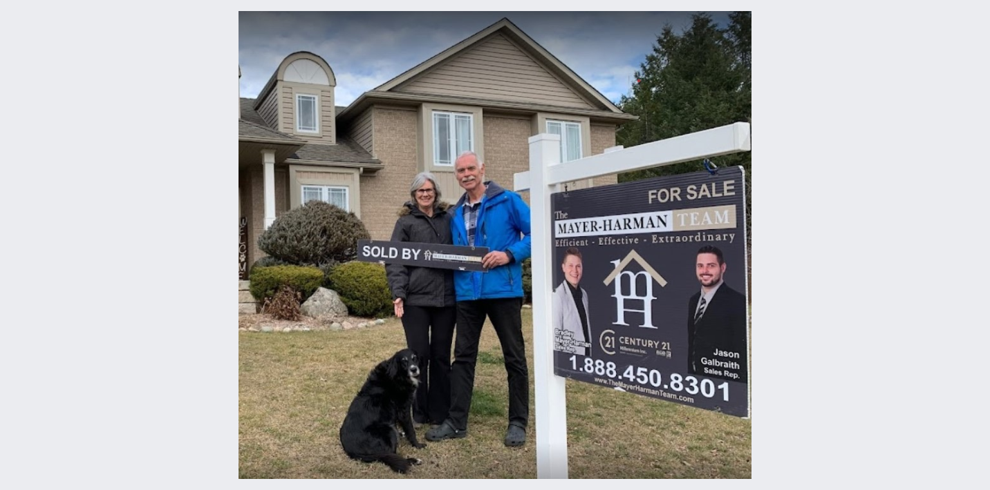 A man and woman are standing in front of a house with a for sale sign.