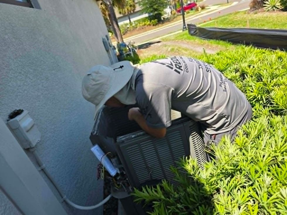 Worker in a sun hat bends over an outdoor HVAC unit beside a house, surrounded by greenery.
