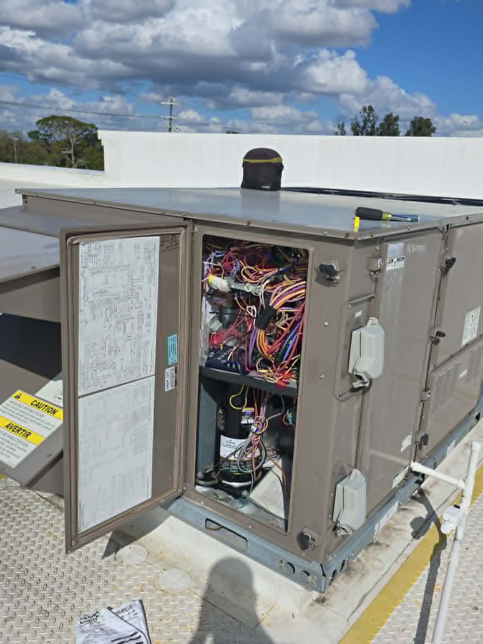 Open electrical control panel on a rooftop HVAC unit with exposed wiring under a cloudy sky
