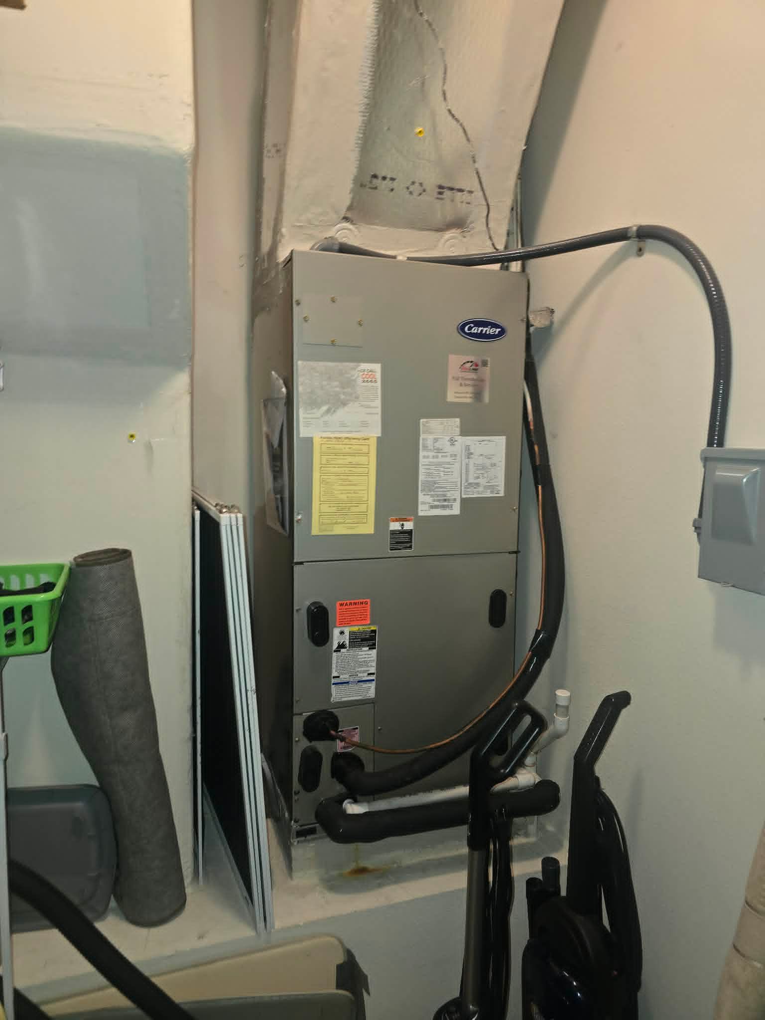 Gray HVAC unit mounted on wall in a utility closet, with black hoses and vacuum equipment nearby