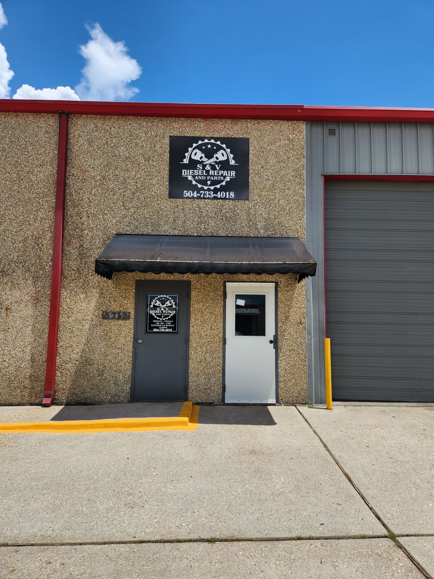 Small industrial building entrance with black awning, gray door, roll-up garage door, and sign above. | S&V Diesel Repair