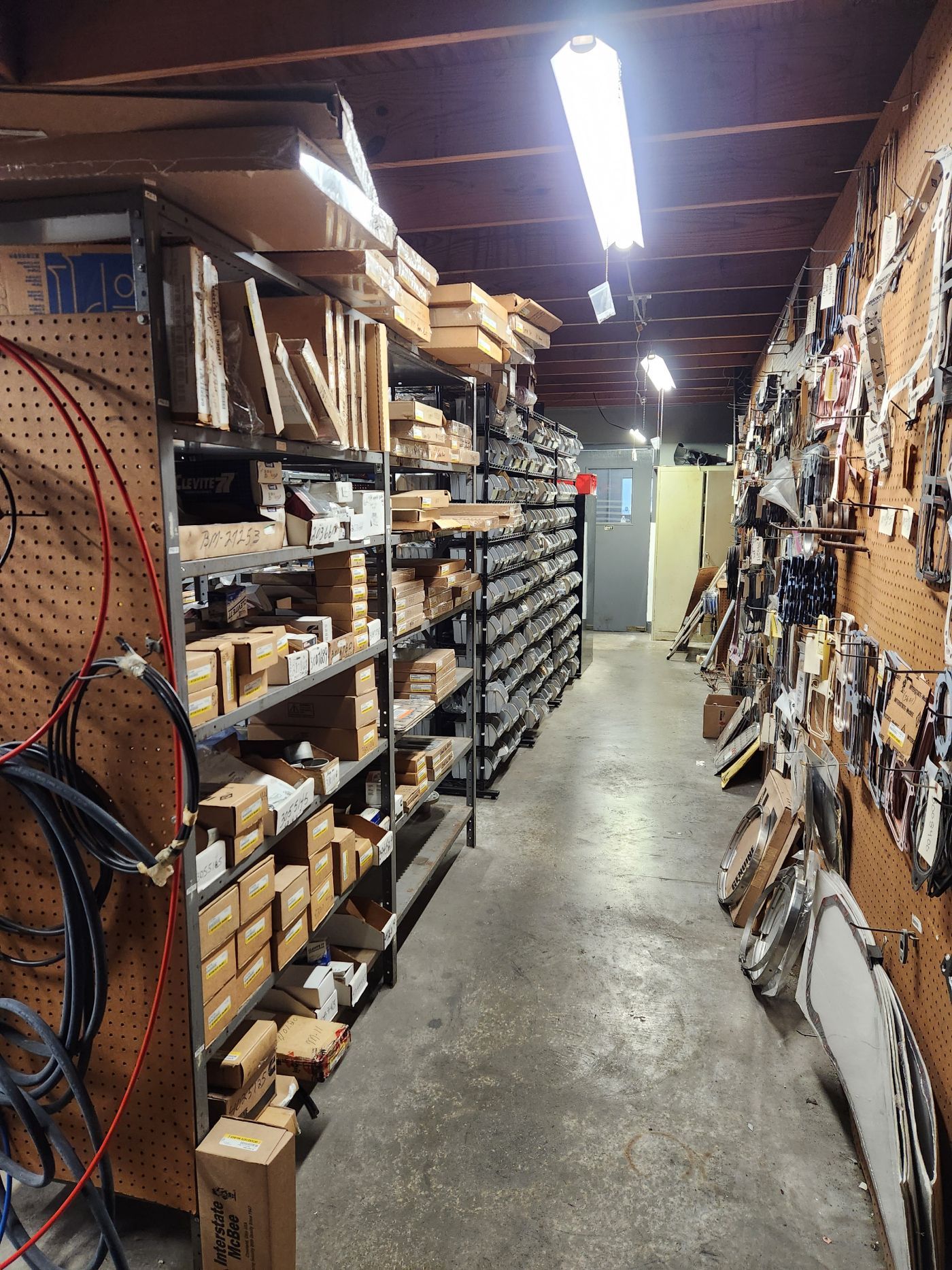 Narrow workshop aisle with shelves of tools, boxes, and hardware under bright fluorescent lights. | S&V Diesel Repair