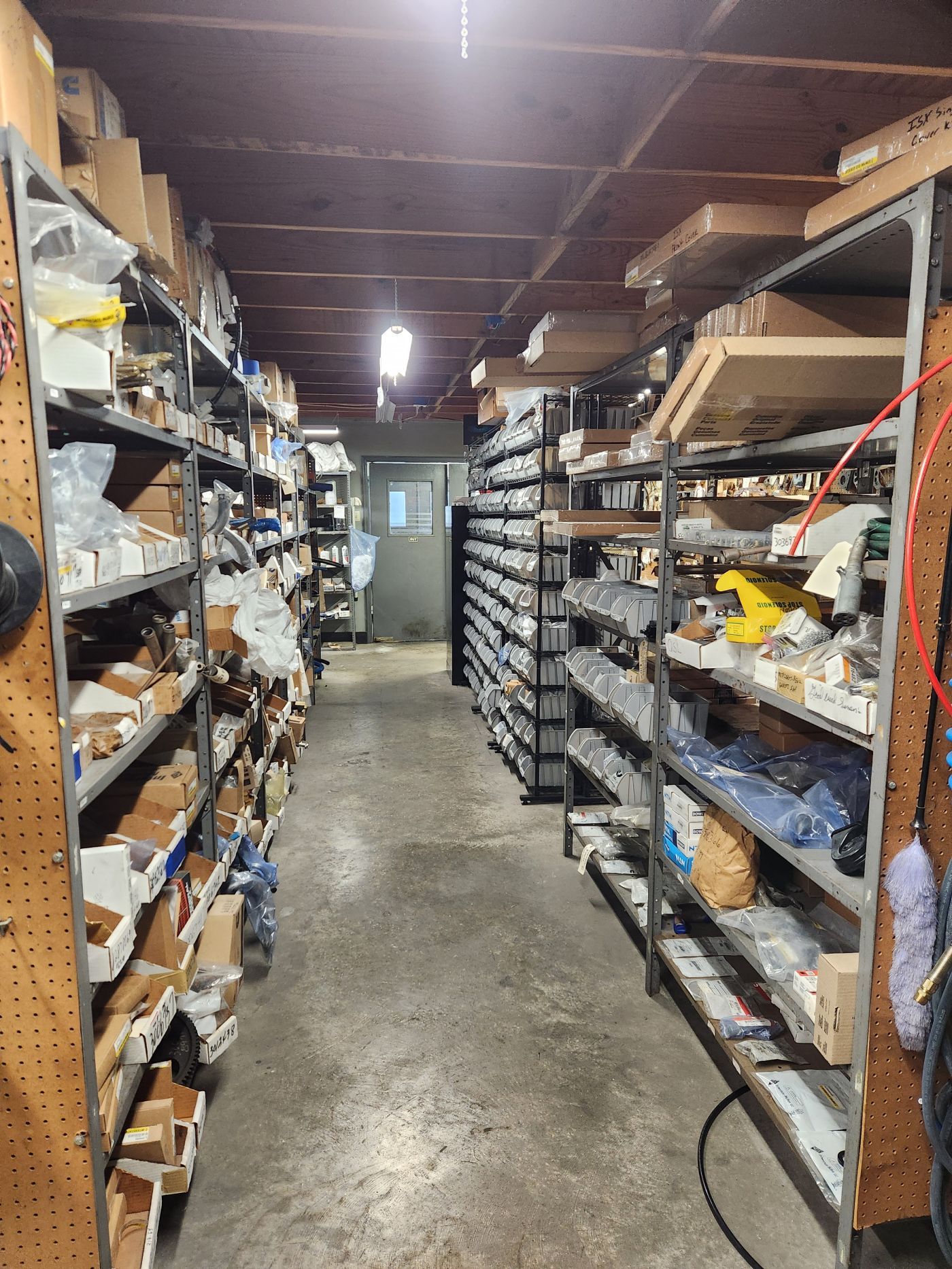 Narrow storeroom aisle with metal shelves packed with boxes, parts, and supplies under overhead lights | S&V Diesel Repair