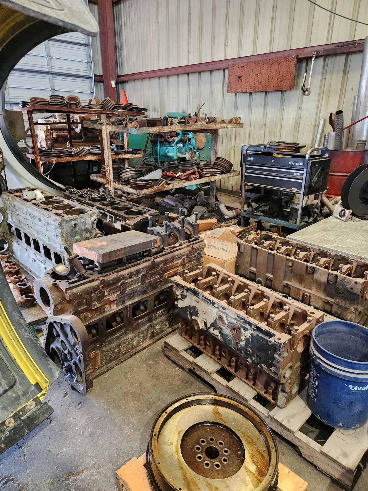 Rusty engine parts and machinery debris stacked in a workshop, with a blue barrel and wheel in the foreground | S&V Diesel Repair