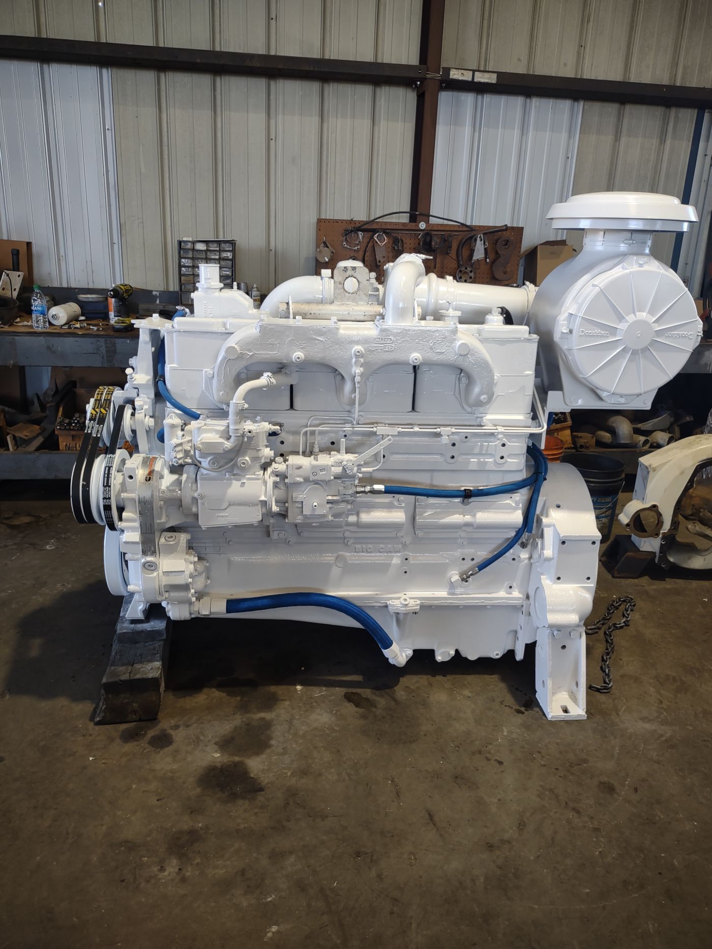 White industrial engine on a pallet in a warehouse workshop, with blue hoses and metal machinery nearby | S&V Diesel Repair
