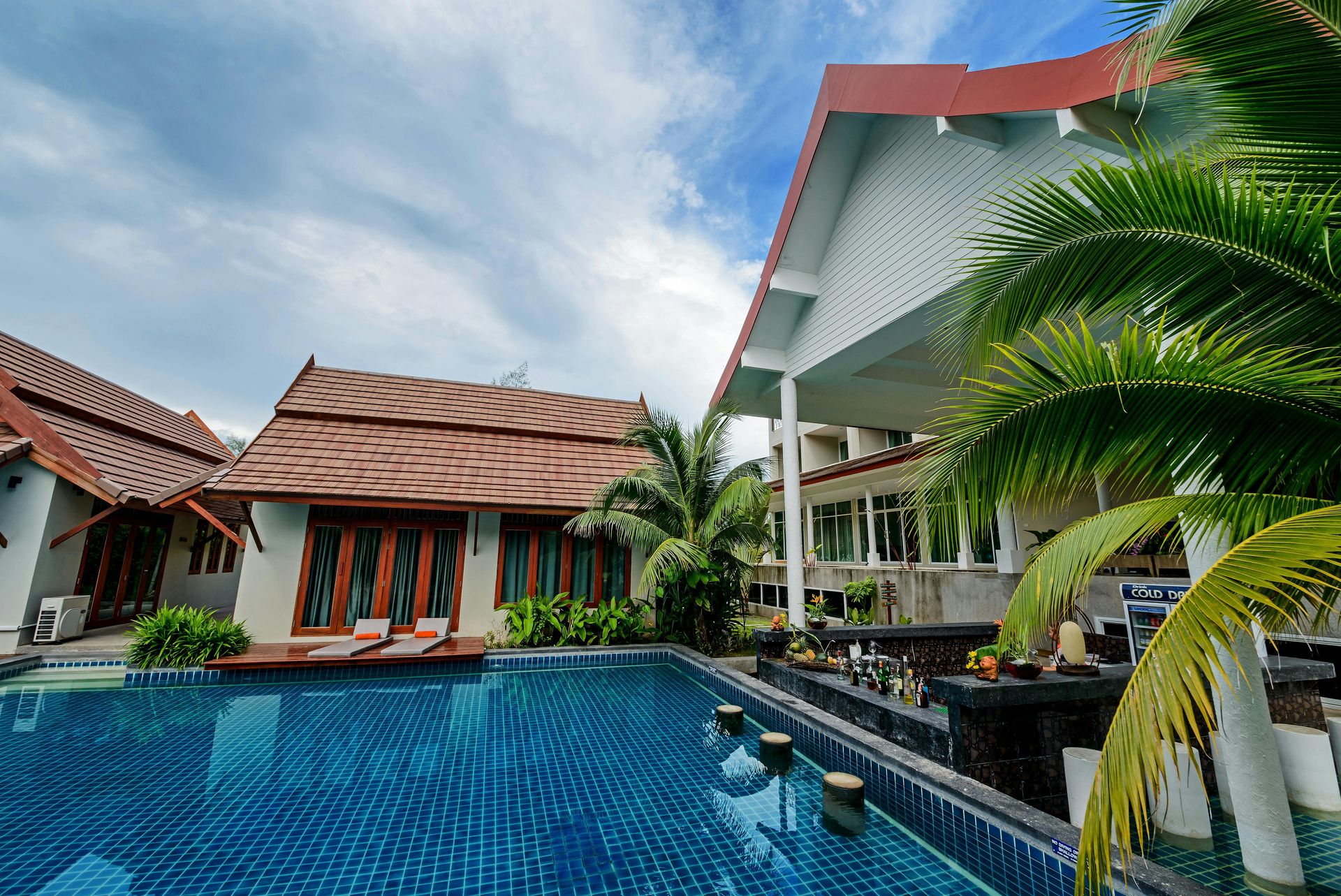 Swimming pool in front of resort buildings with brown tiled roofs. Palm trees, blue sky.