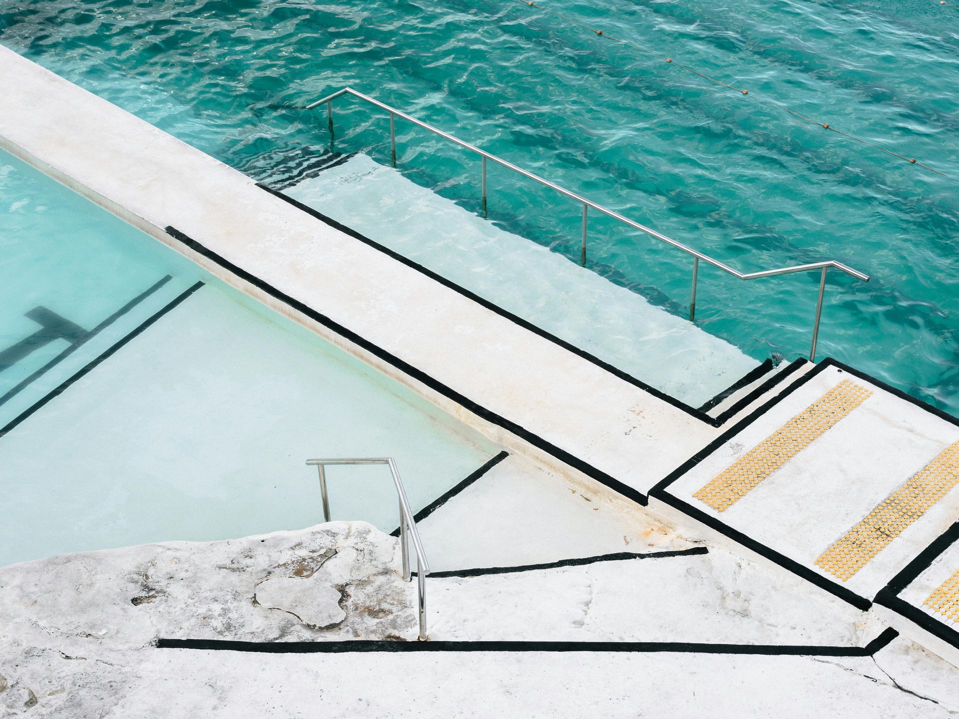White concrete steps leading into a turquoise ocean pool. Black and yellow markings enhance the steps.