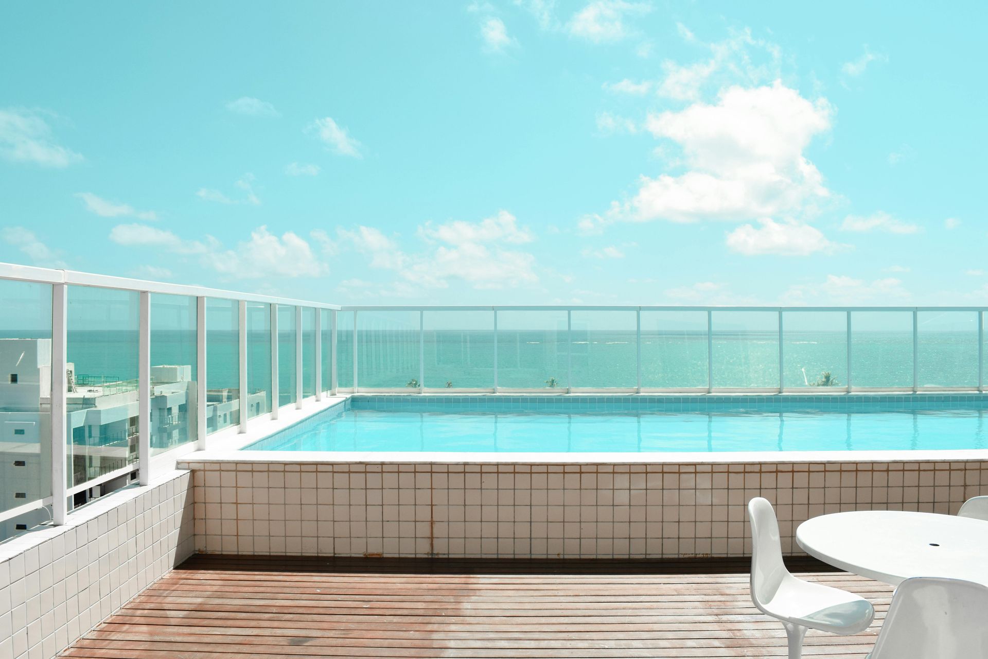 Rooftop pool overlooking the ocean, with white railing and chairs, on a sunny day.