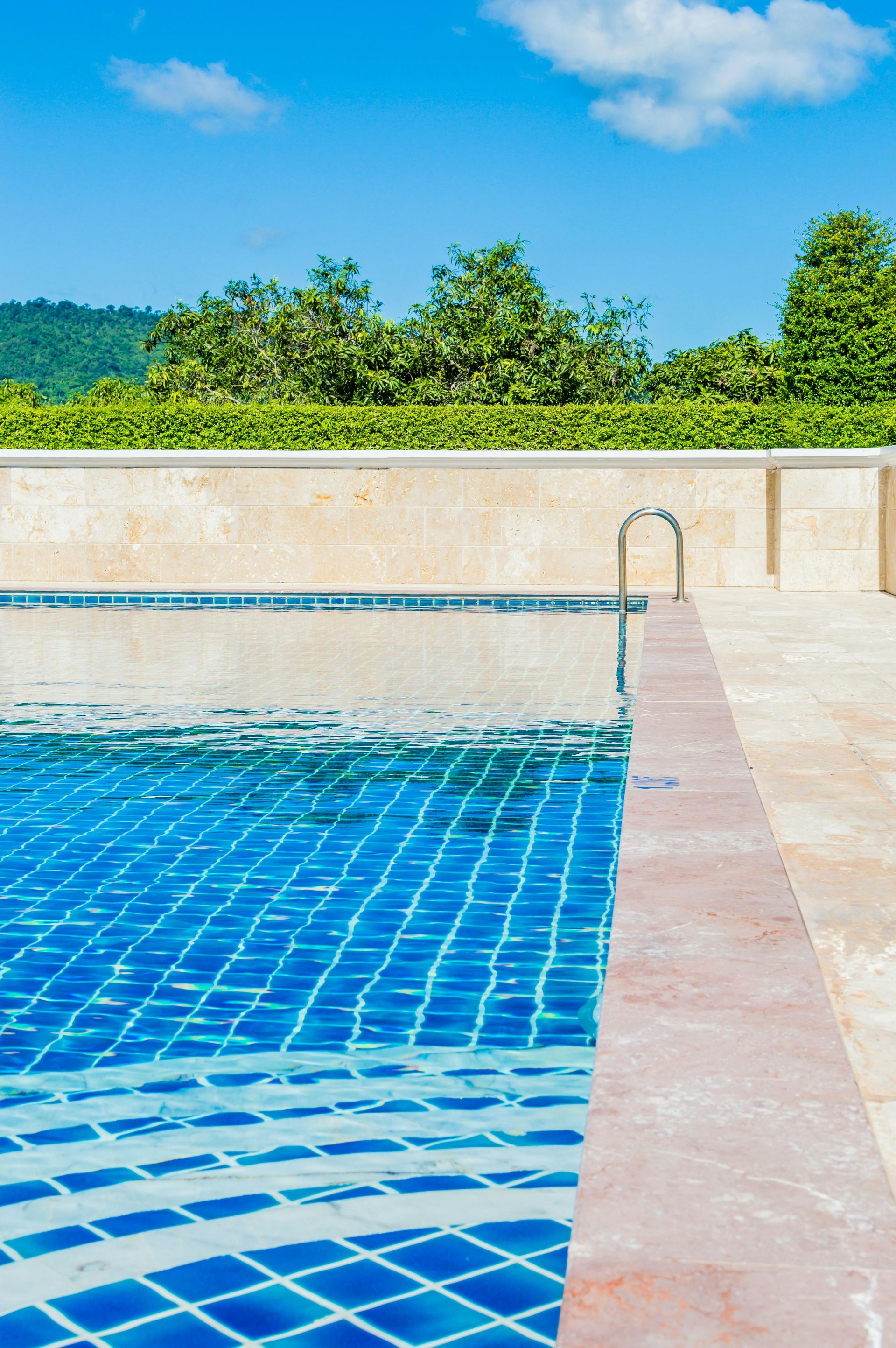 Blue-tiled swimming pool with ladder, beige border, under blue sky, with green foliage in background.