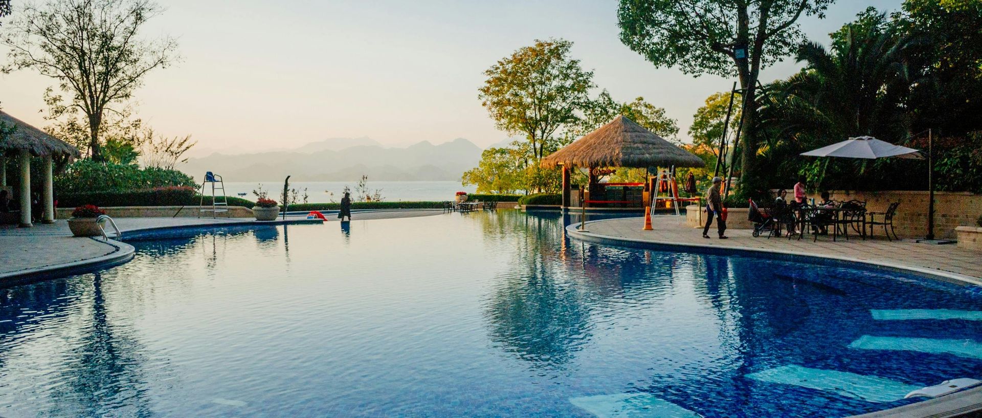 Infinity pool overlooking a calm body of water, with a gazebo and people sitting at tables.