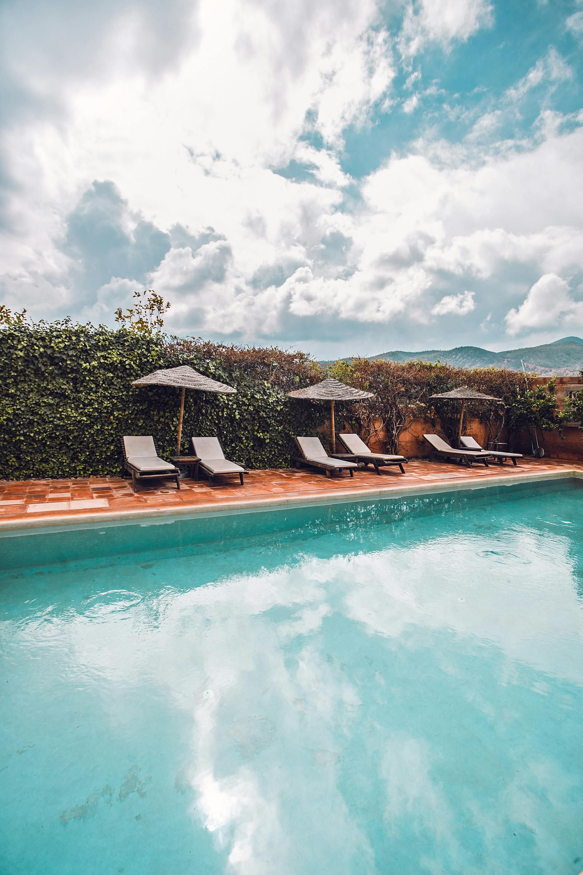 Swimming pool with lounge chairs, umbrellas, and cloudy sky reflecting in water.