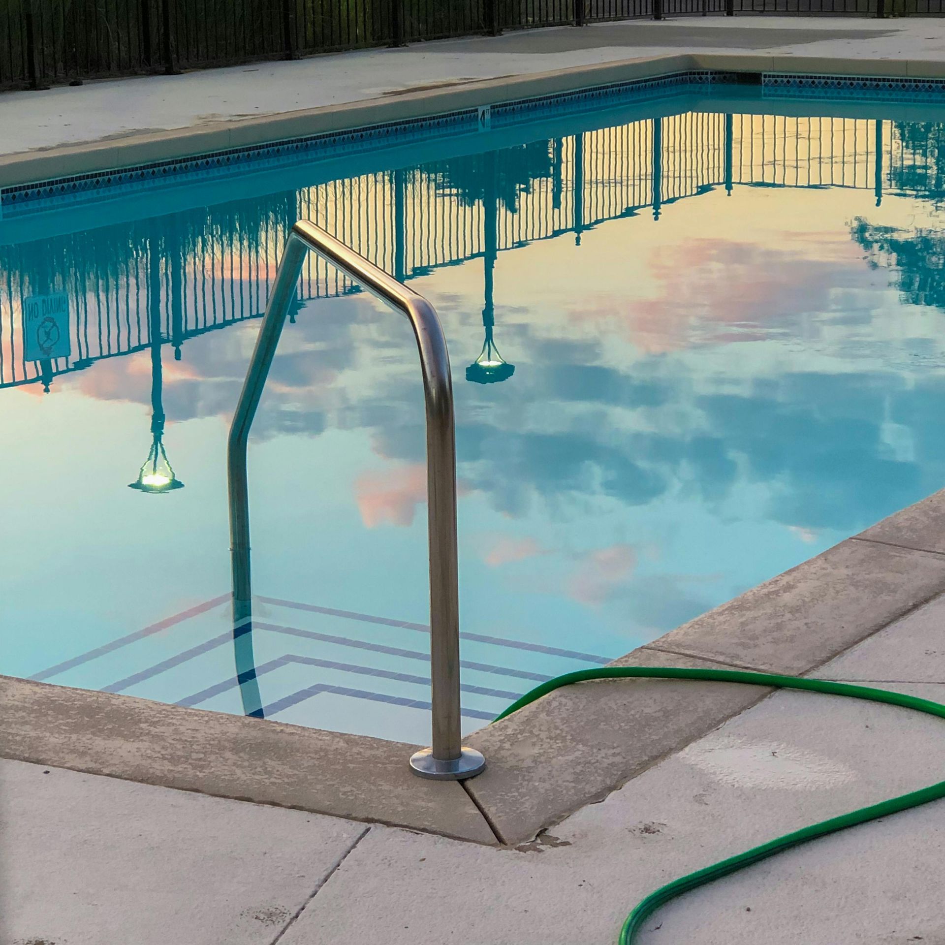 Swimming pool with steps, handrail, and reflections of sky and fence. Green hose visible.