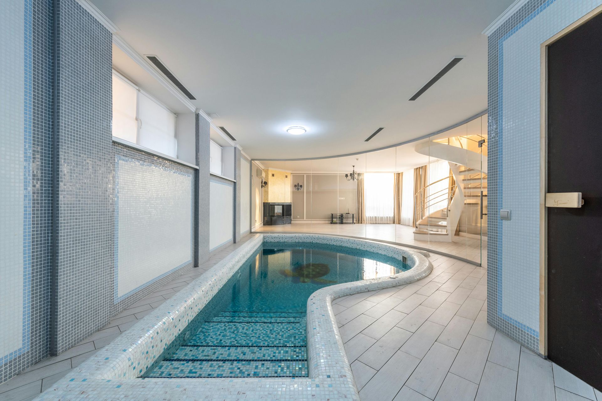 Indoor swimming pool with white tile surround, curved shape, and blue water. White walls and wood flooring.