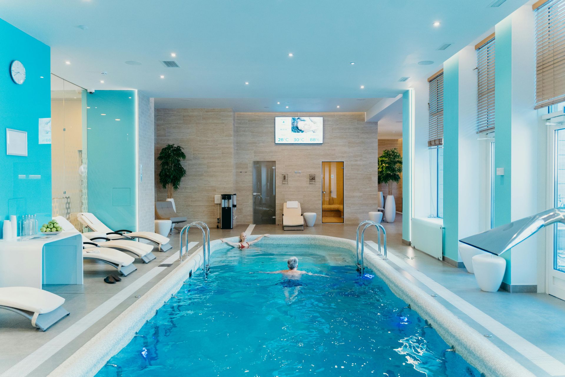 Indoor pool with swimmers, lounge chairs, and spa-like decor; blue and neutral tones.