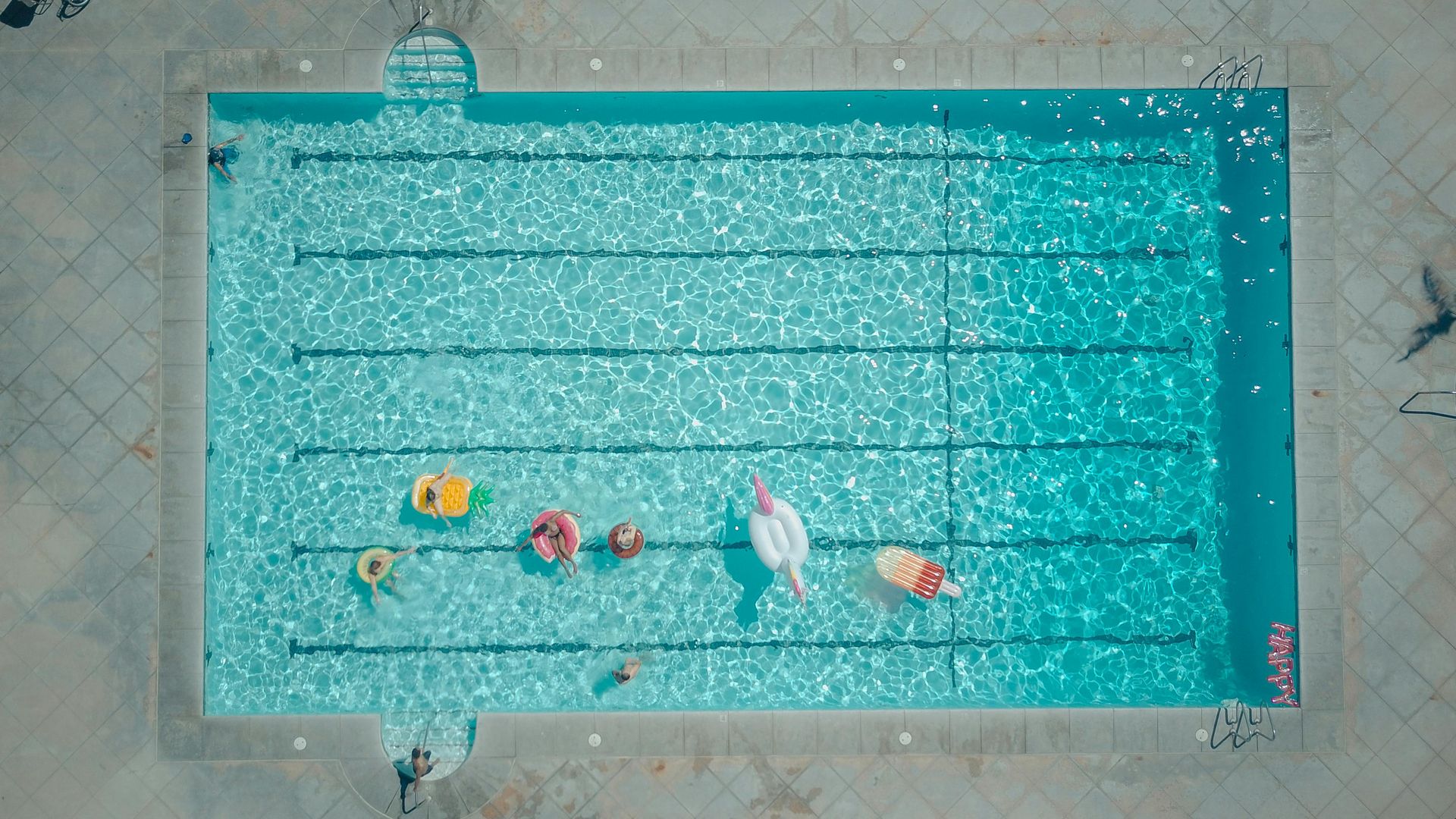 Overhead view of a swimming pool with several inflatable floats; water is blue and rippled.