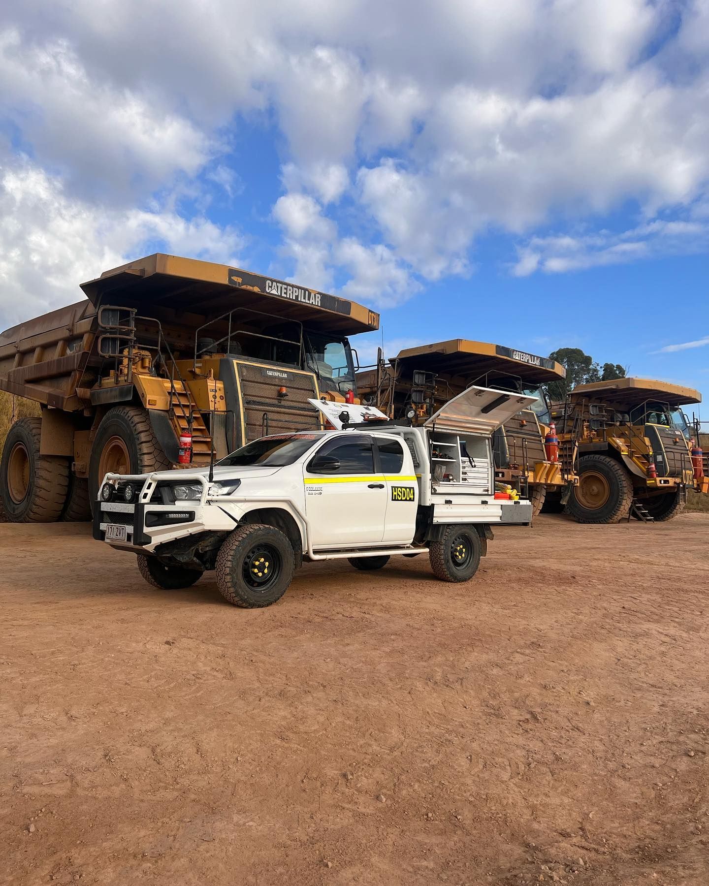 A white utility truck parked between two large Caterpillar mining haul trucks on a dirt lot under a cloudy blue sky.