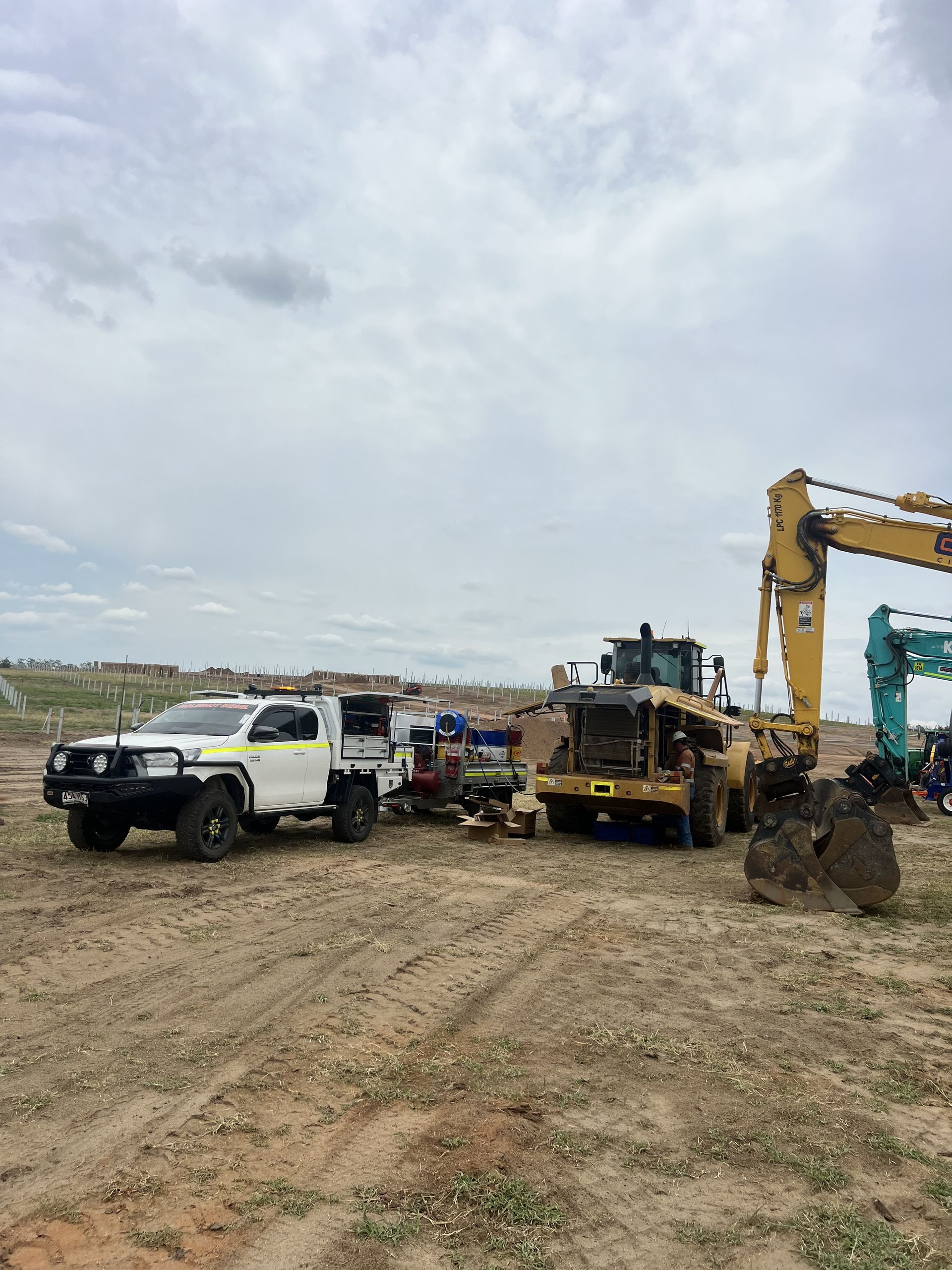 A white service truck parked next to yellow and teal construction excavators in an open, dirt field under a cloudy sky.