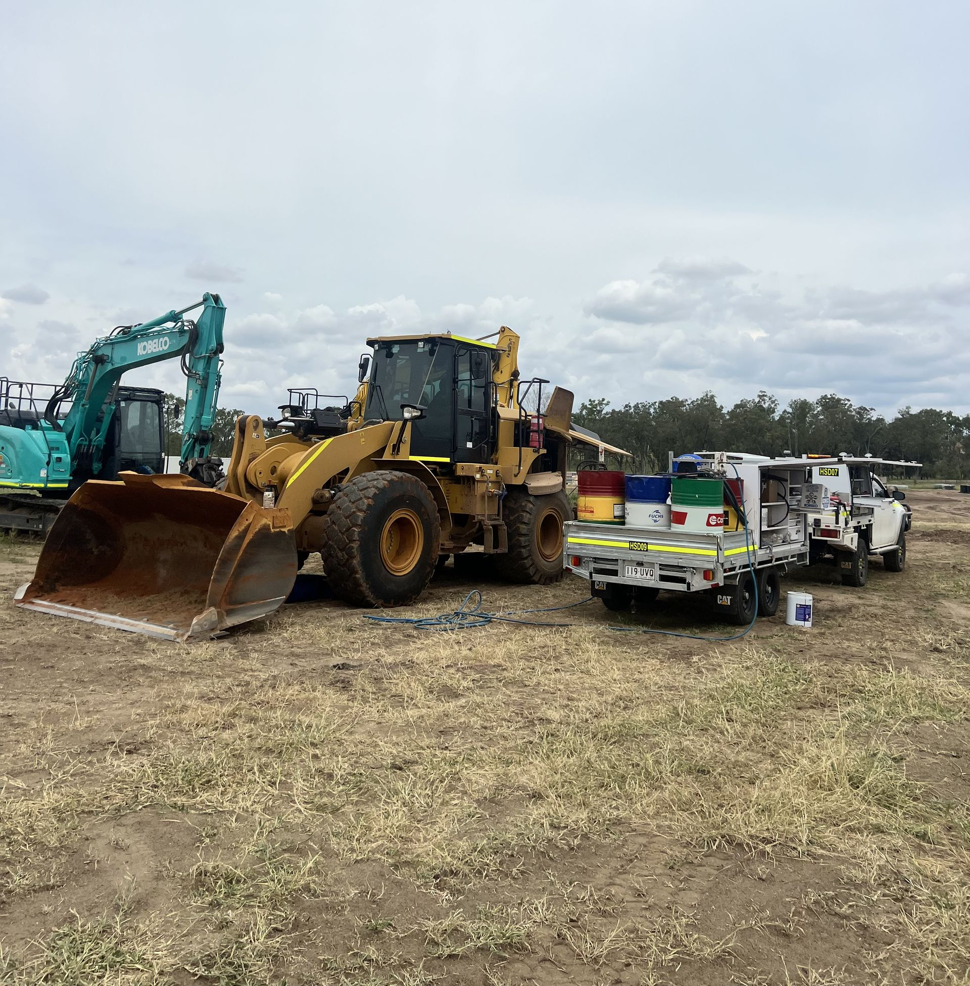A yellow wheel loader and a teal excavator parked on a dirt field with service trucks carrying fuel drums nearby.