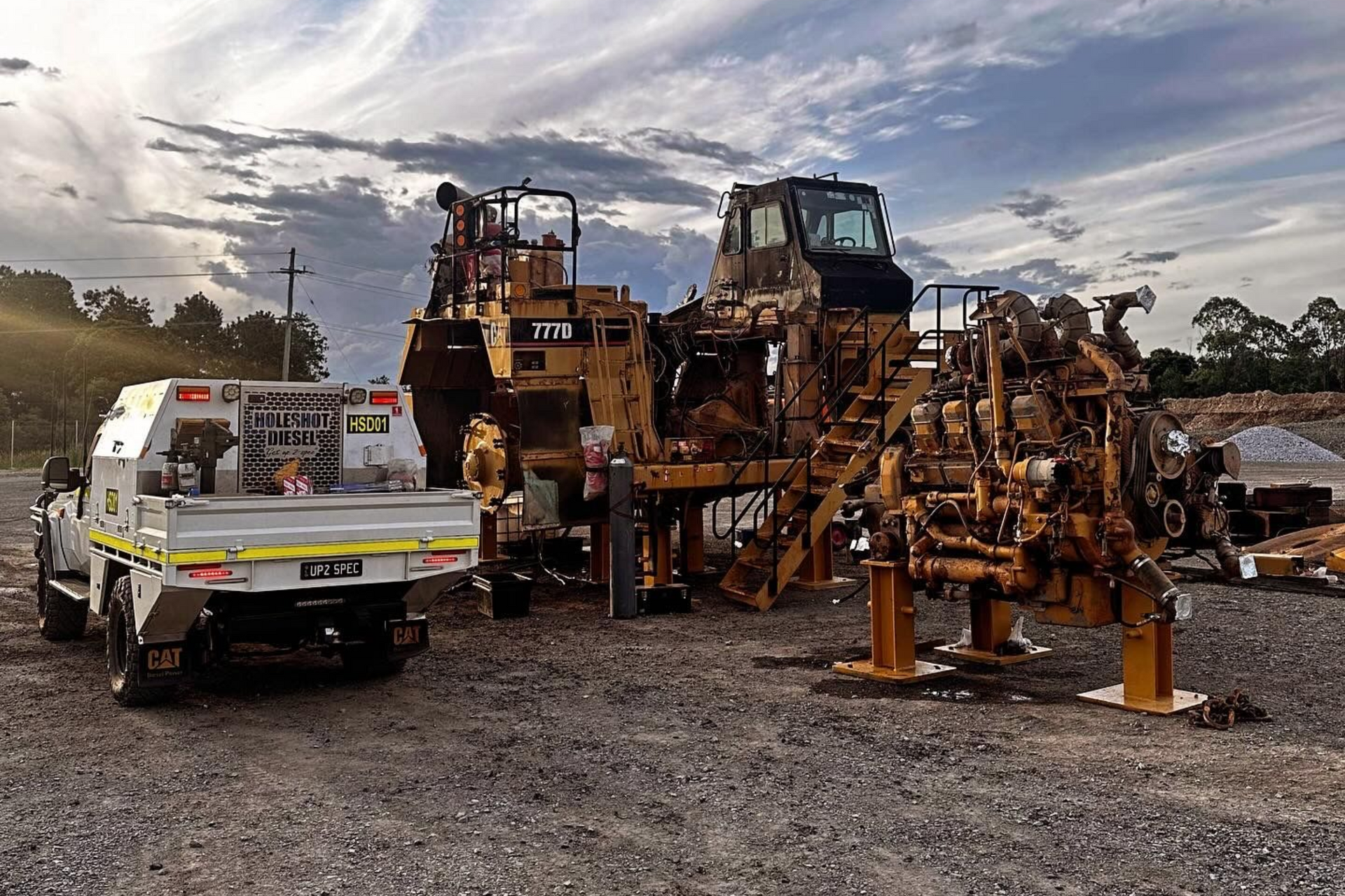 A utility truck parked next to a large piece of disassembled yellow construction machinery on a gravel lot at dusk.