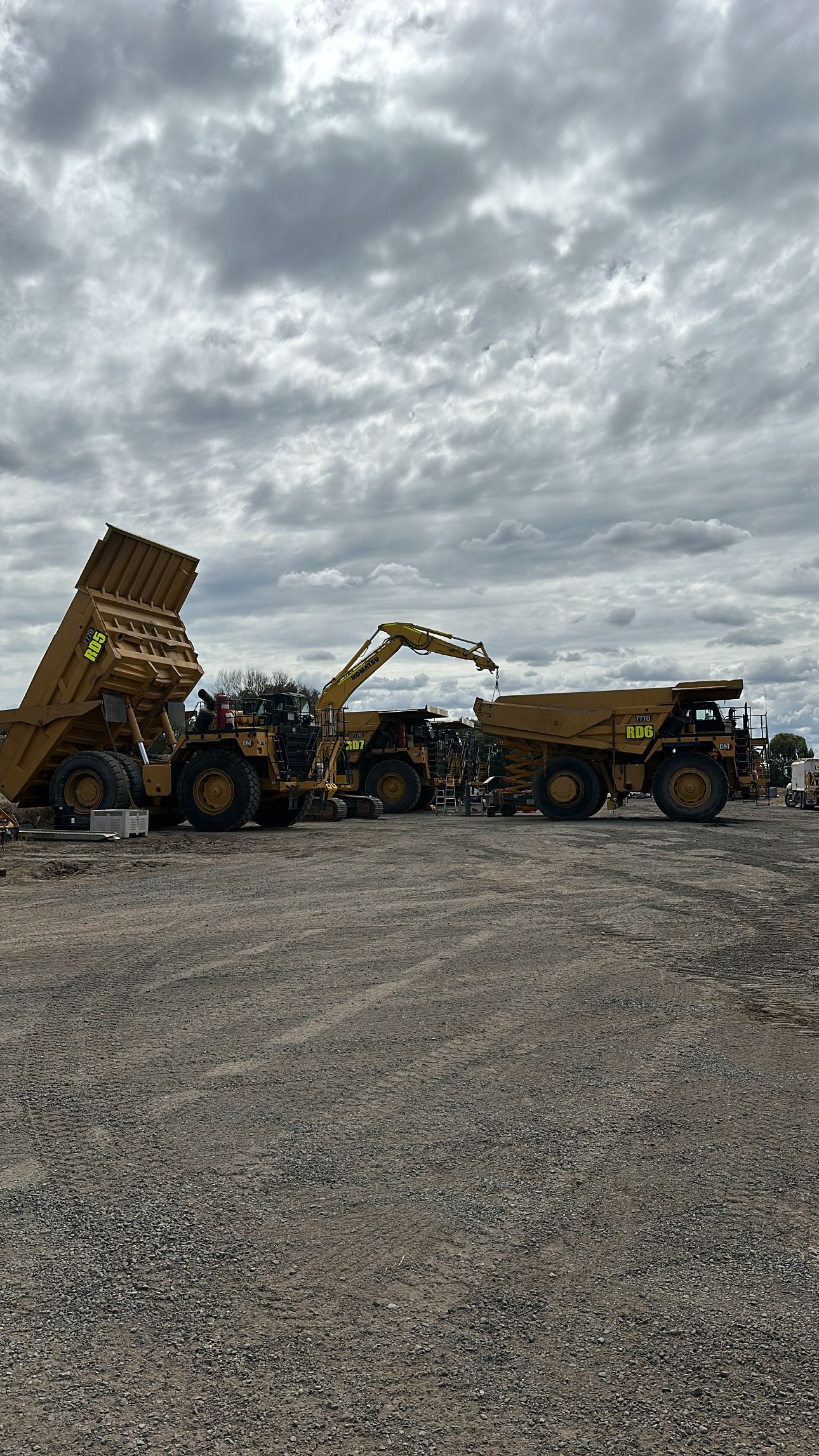 Yellow construction vehicles at a work site under a cloudy sky. One dump truck has its bed raised.