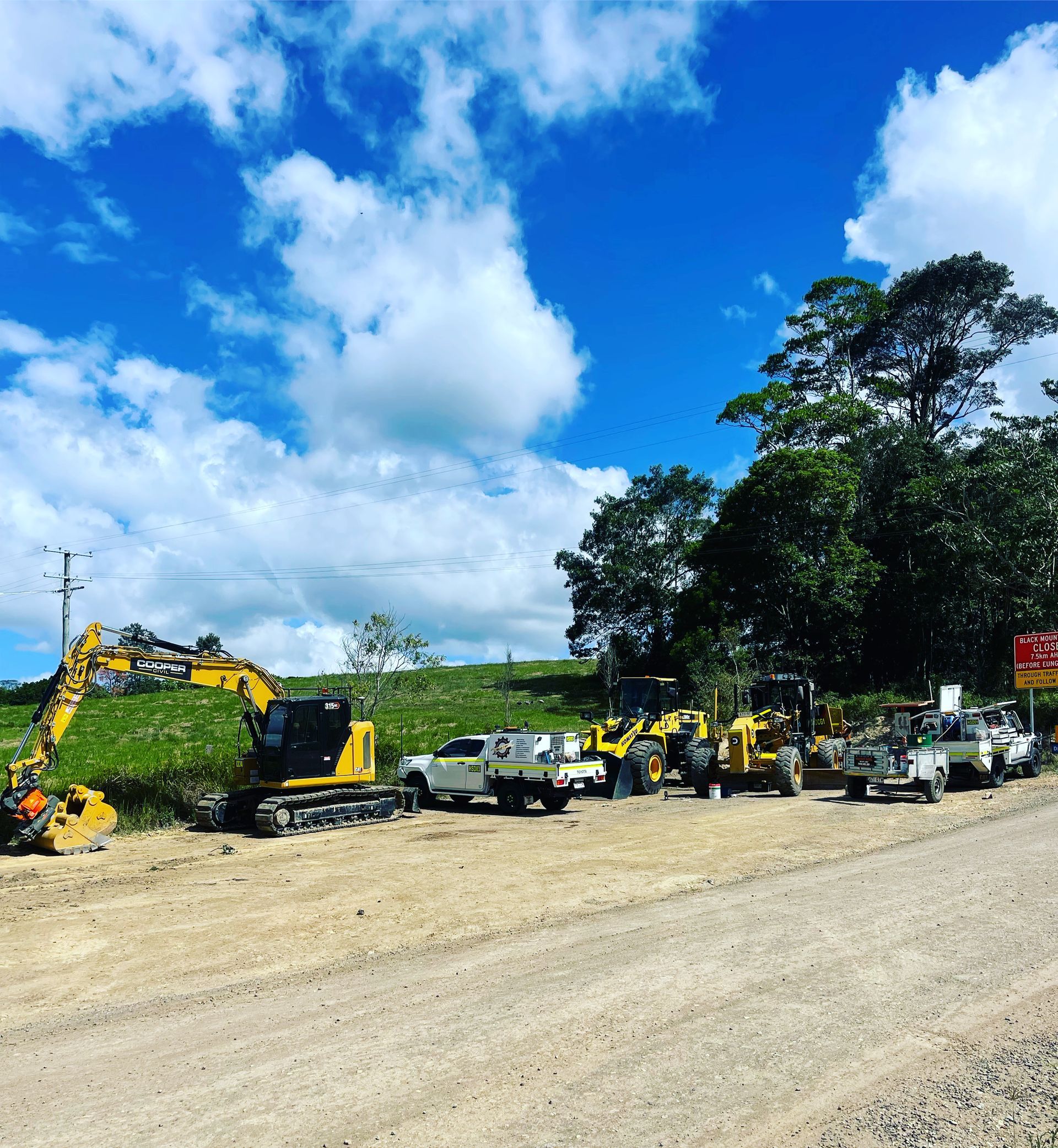 A line of yellow construction equipment and parked trucks on a dirt roadside under a bright, partly cloudy sky.