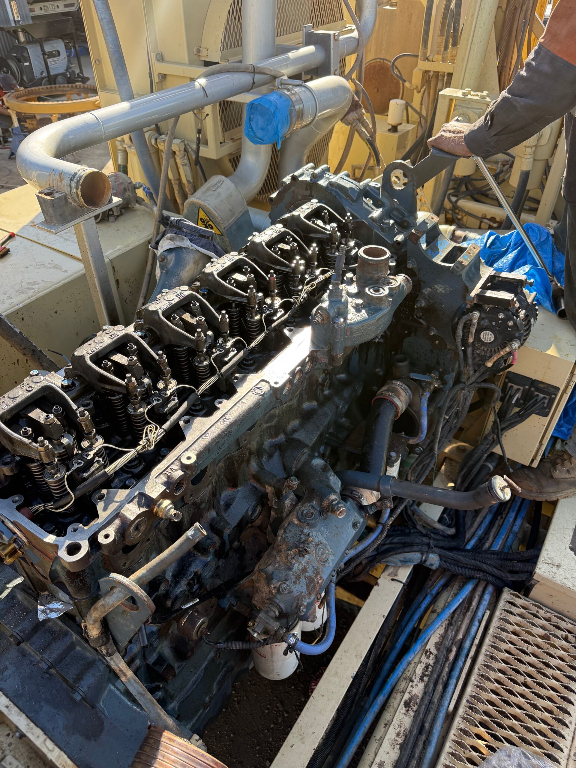 A large, dark industrial diesel engine with its valve cover removed, undergoing maintenance on a heavy machinery site.