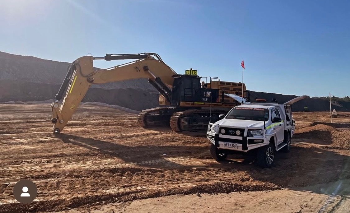 A yellow excavator parked next to a white pickup truck on a dirt worksite under a clear blue sky.