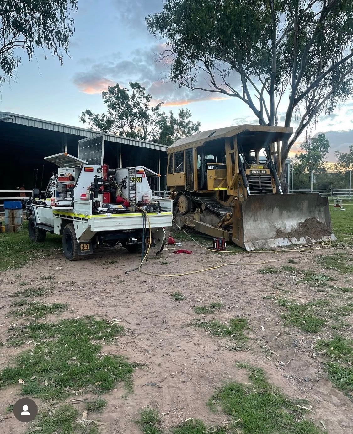 A service truck parked beside a large yellow bulldozer in an outdoor area near a shed at dusk.