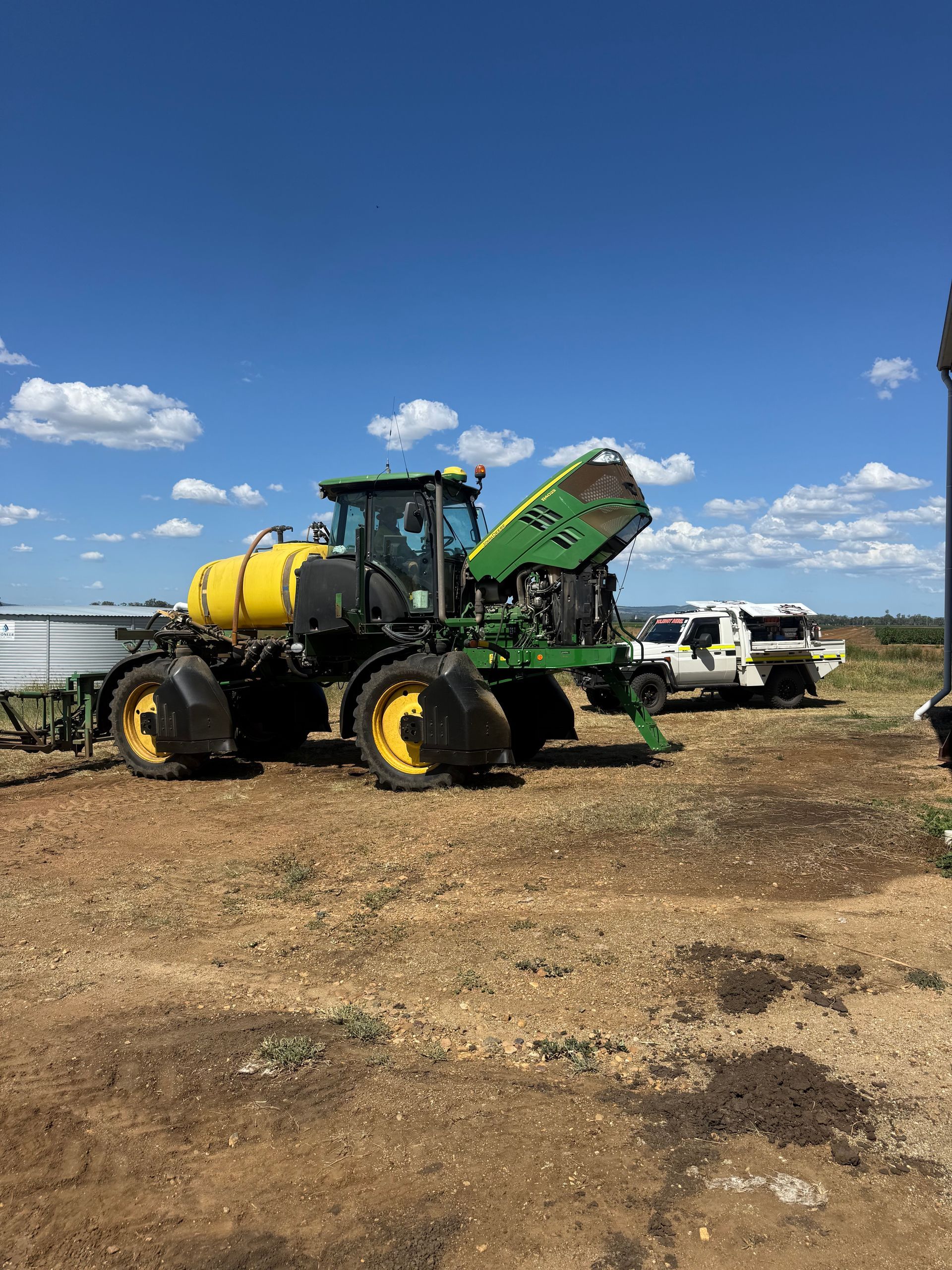 A green John Deere self-propelled sprayer with its hood open, parked in a dirt field with a white utility vehicle nearby.