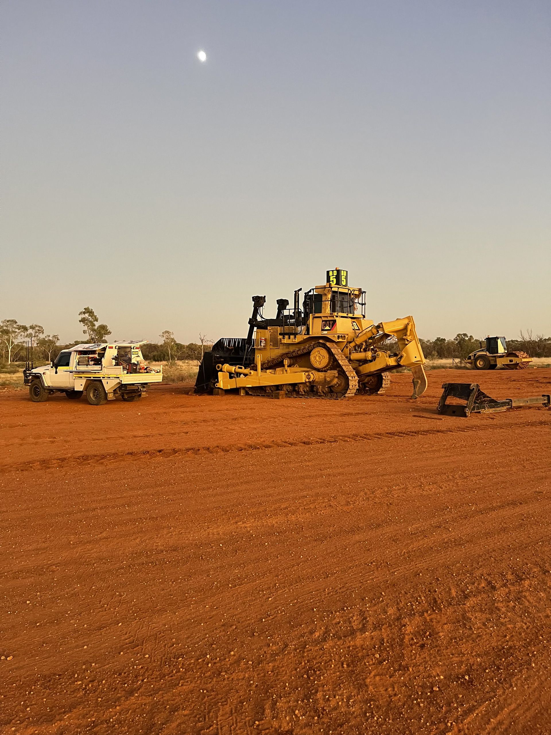 A yellow bulldozer and a white utility vehicle work on an expansive, barren red dirt landscape under a bright sky.