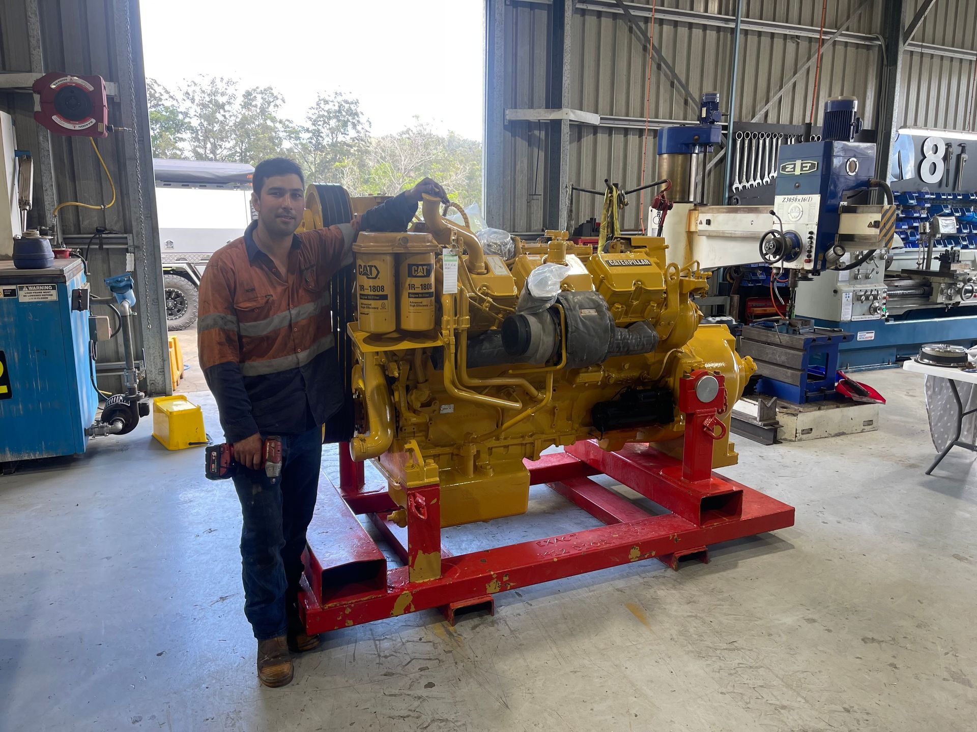 A technician stands beside a large yellow engine mounted on a red metal frame in a warehouse workshop.