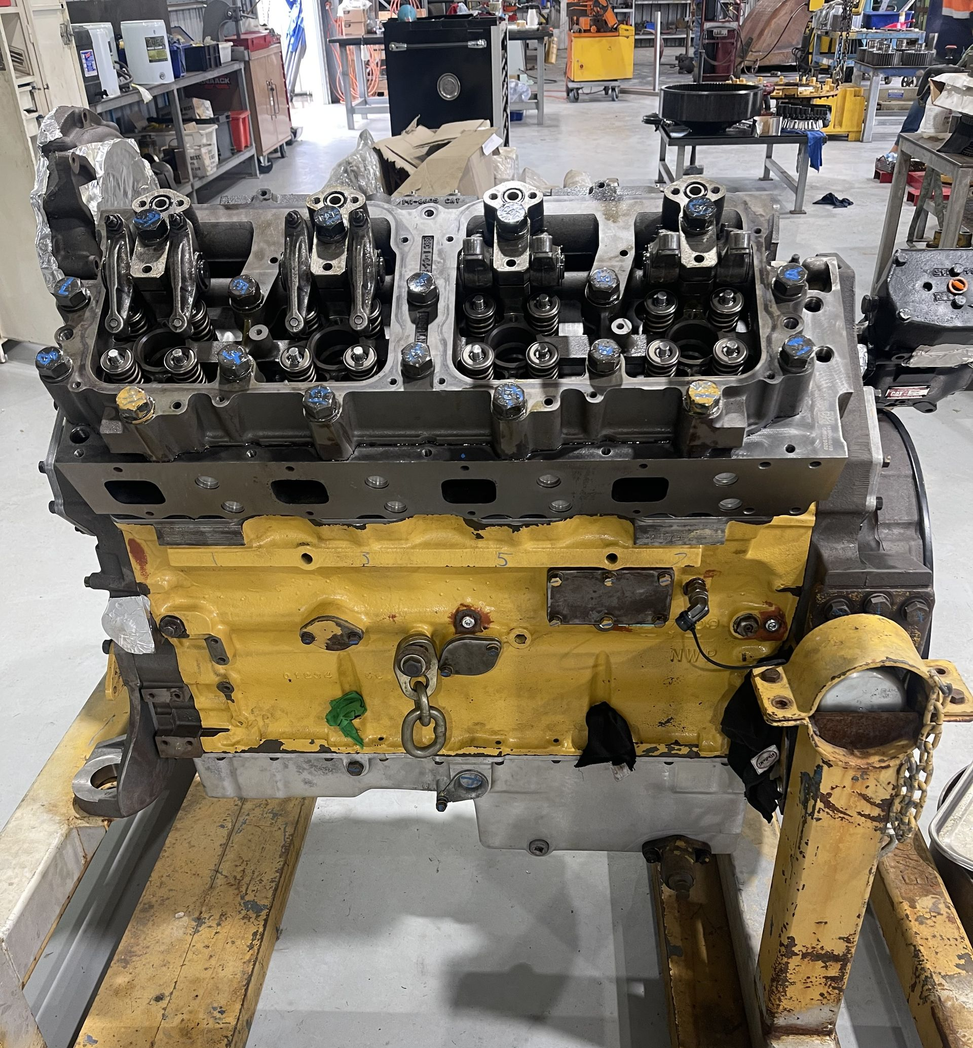 A yellow Caterpillar diesel engine block with an exposed cylinder head sits on a work stand in a mechanic's shop.