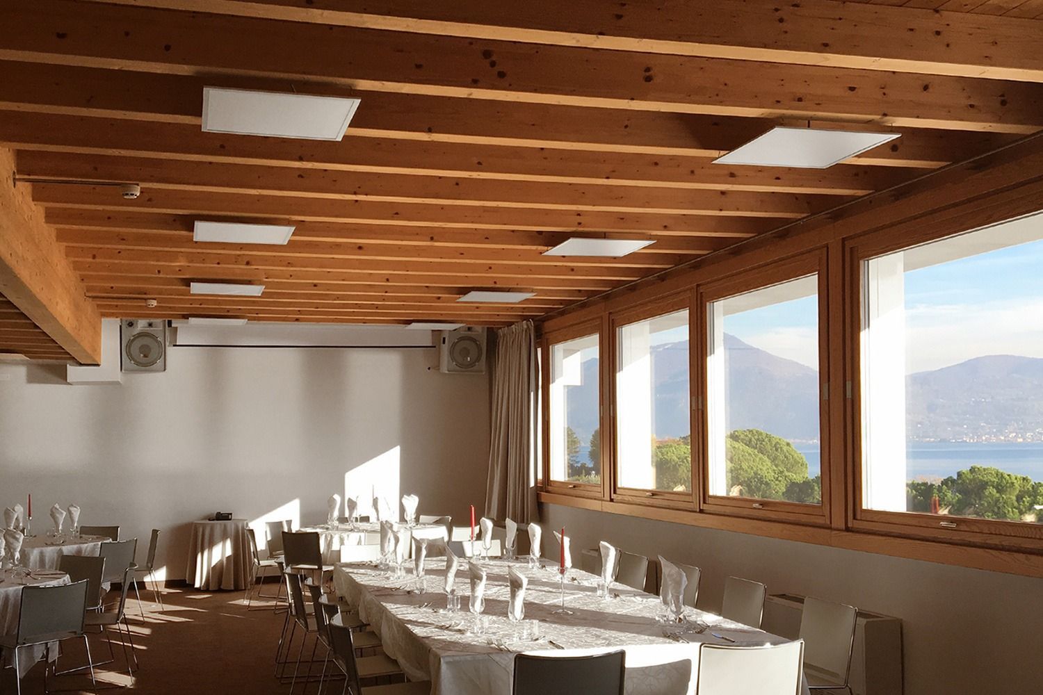 Camera con travi in legno sul soffitto, ampie finestre con vista sul lago e sulle montagne e tavoli apparecchiati per i pasti.