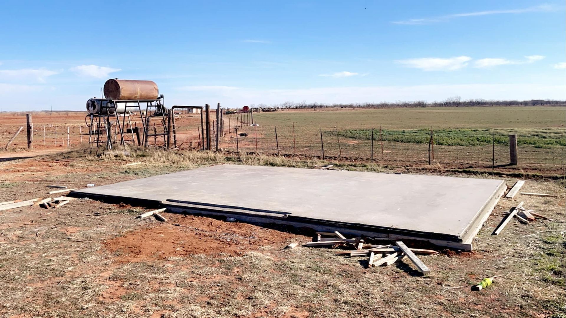 Concrete slab in a field with a water tank and fence in the background, under a blue sky.