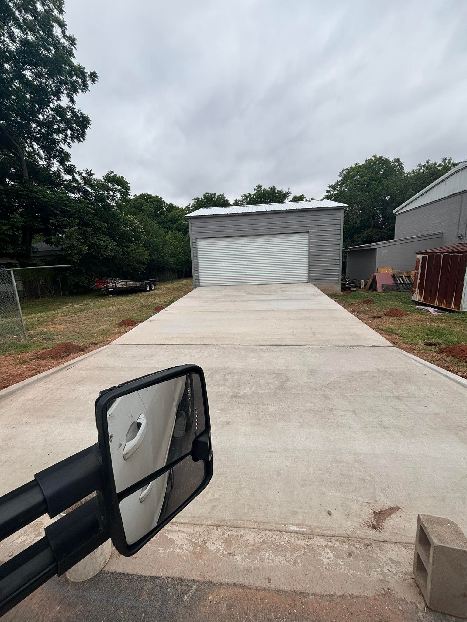 Concrete driveway leads to a metal garage with a closed door, cloudy sky. Side mirror in the foreground.