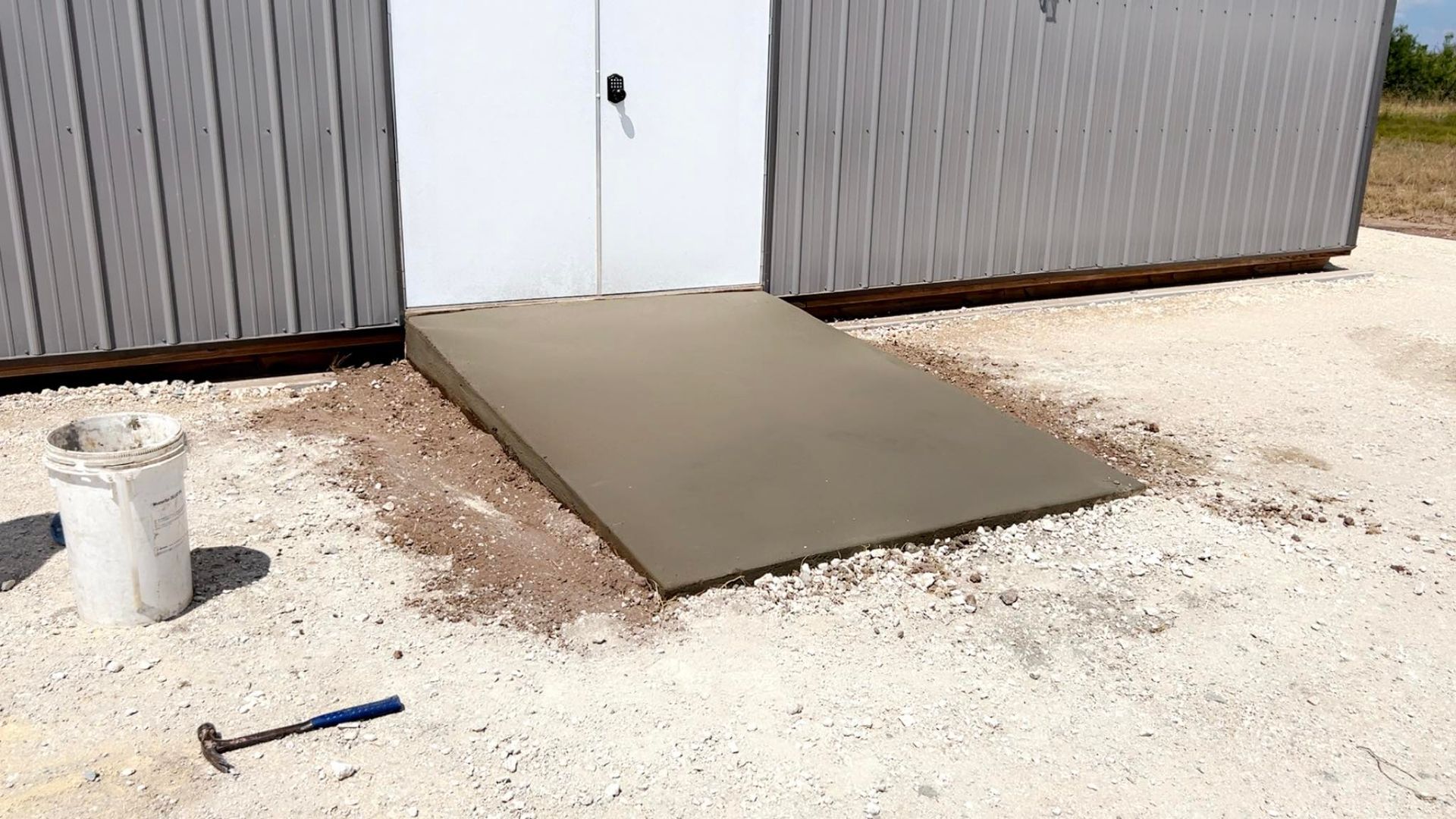 Concrete ramp leading to a shed door, set on gravel. Hammer and bucket of concrete mix nearby.